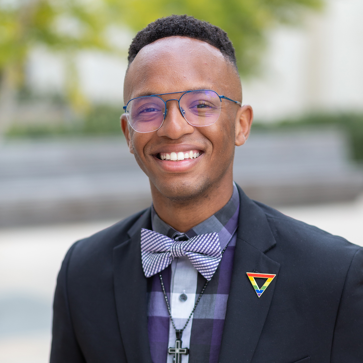 Black male wearing glasses, suit jacket, checkered shirt, bowtie, pride pin, cross necklace